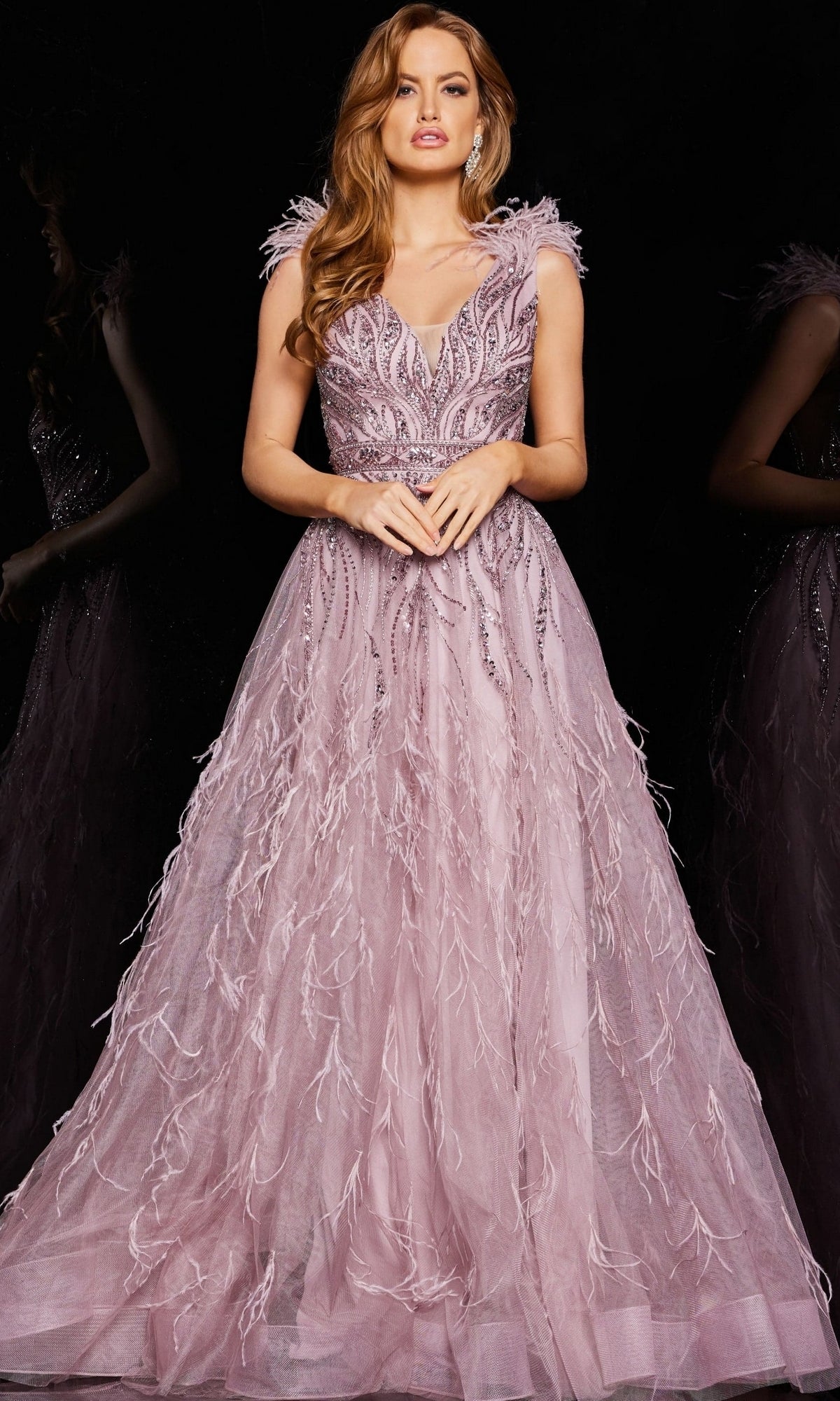 A woman stands against a black background wearing the Jovani Feathered Long Pink Prom Dress 24000, a floor-length gown adorned with feather and sequin embellishments.